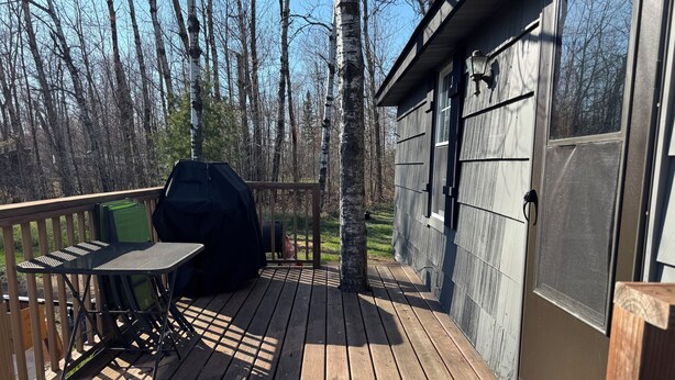 A wooden deck with a covered grill, a table, and chairs. - Charming cabin in delightful Duquette with AC near miles of ATV trails. (Kerrick)