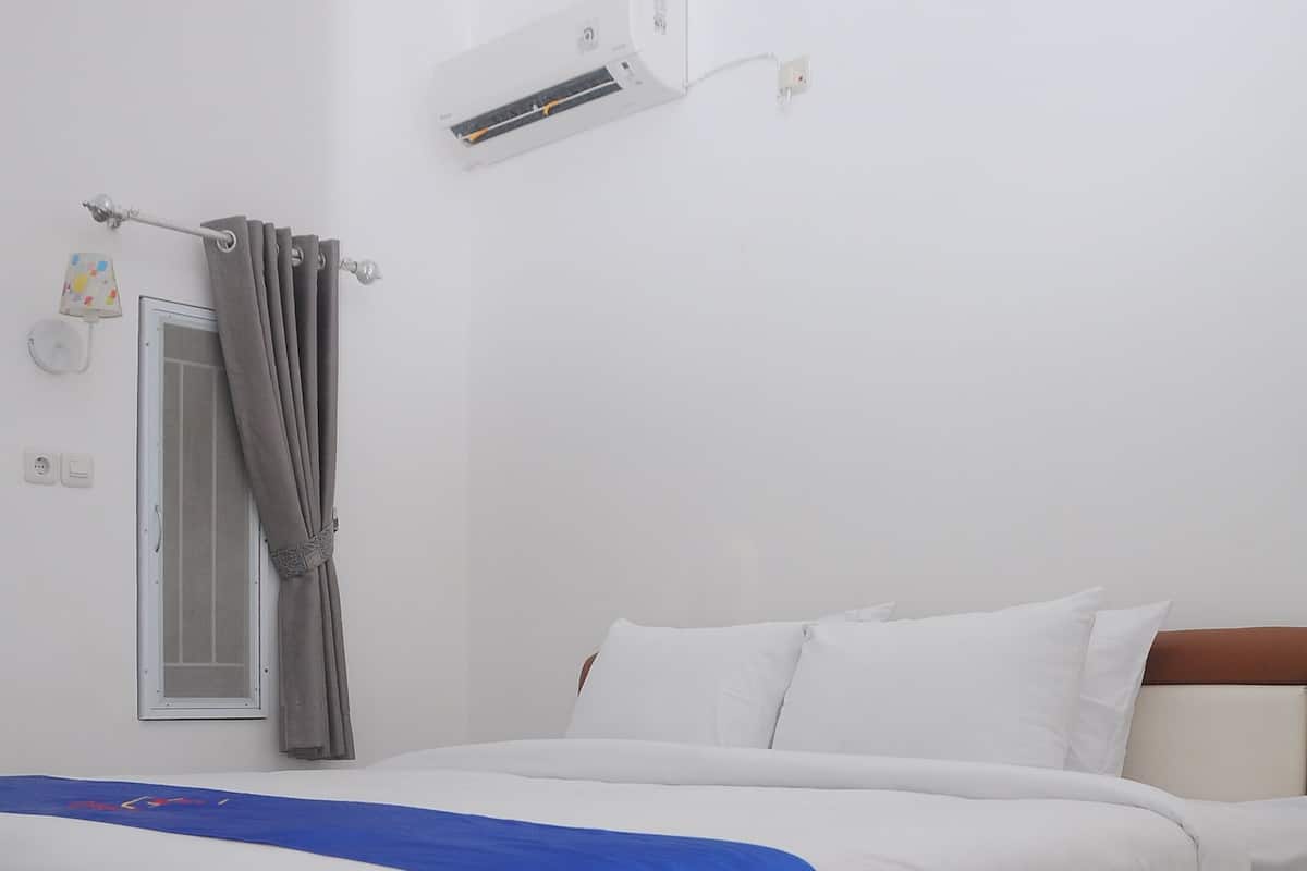Een hotelkamer met een bed, witte muren, een raam met gordijn, een airconditioning en een klein decoratief element aan de muur.