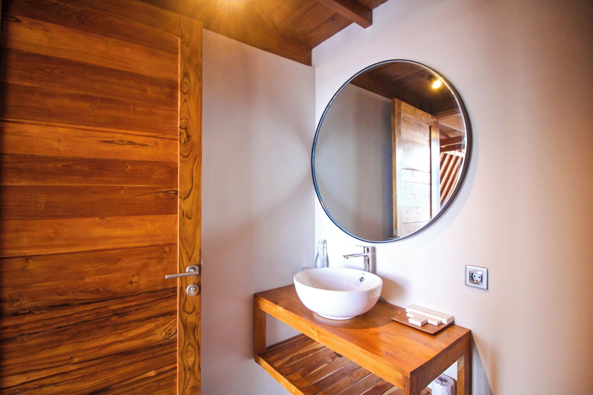 A wooden bathroom with a round mirror, a white sink, and a wooden shelf.