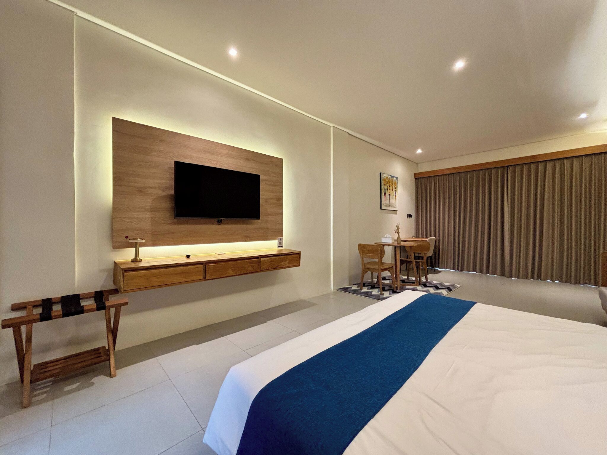 A modern hotel room with a large bed, a flat-screen TV mounted on a wooden panel, a small desk with a chair, and a dining area with a table and chairs.