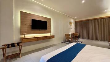 A modern hotel room with a large bed, a flat-screen TV mounted on a wooden panel, a small desk with a chair, and a dining area with a table and chairs.
