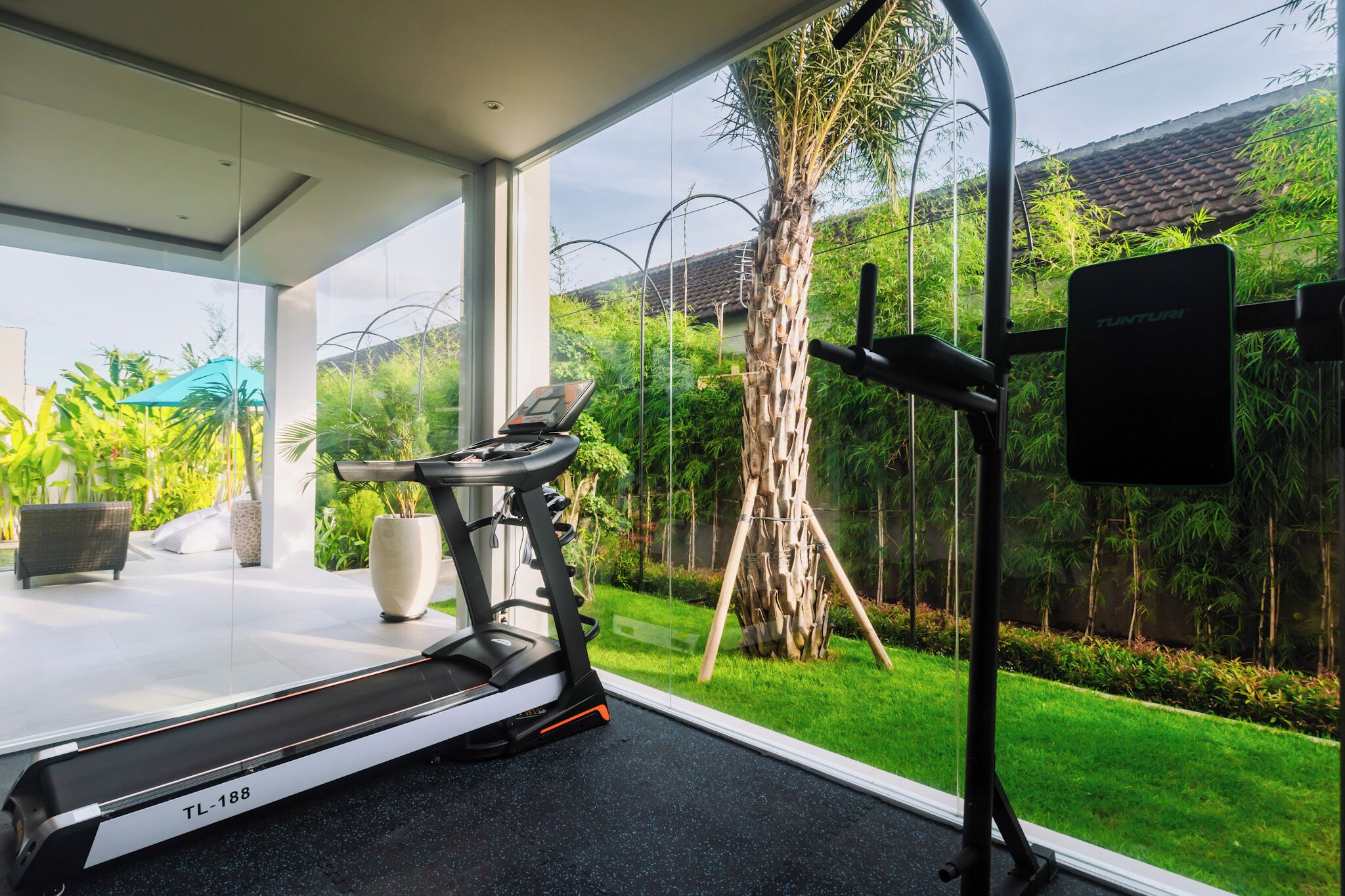 Et moderne fitnessrum med løbebånd, træningsudstyr og et stort vindue med udsigt til en frodig have.