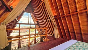 A room with a sloped wooden ceiling, a bed with a patterned bedspread, and a view of the outside through a large window. - Campbar 4 (Munggu)