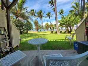 Une terrasse avec une table ronde et deux chaises, une petite étagère avec quelques objets, et une vue sur un espace verdoyant agrémenté de palmiers et d’une étendue d’eau.