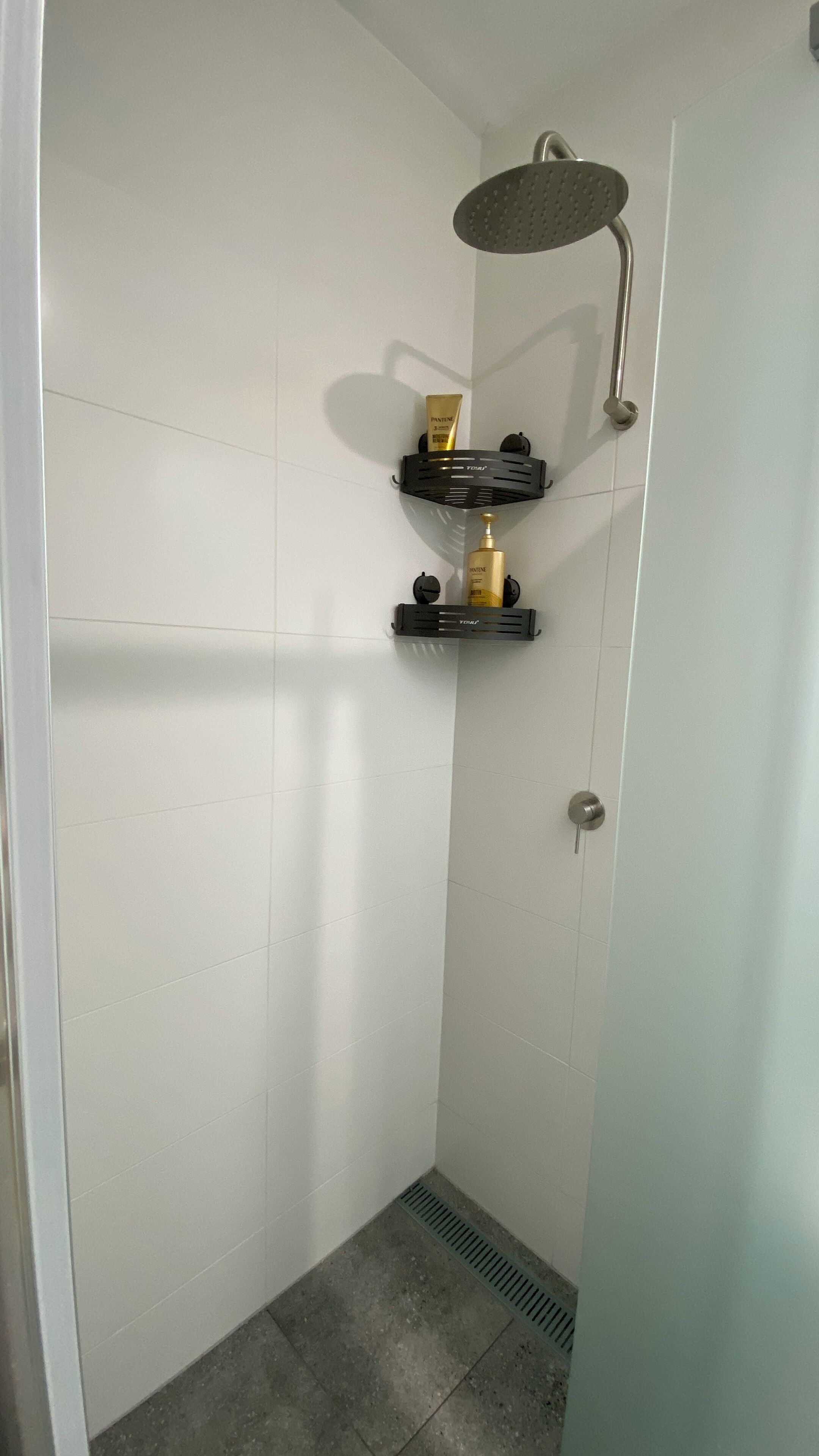 Un pequeño box de ducha con azulejos blancos, un cabezal de ducha y un estante negro con artículos de aseo.