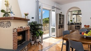 A dining area with a wooden table, chairs, and a bowl of fruit. There is a fireplace, a kitchen visible through an open door, and a seating area with a sofa and armchairs. - Villa Marcella with private pool and sea views (Massa Lubrense)