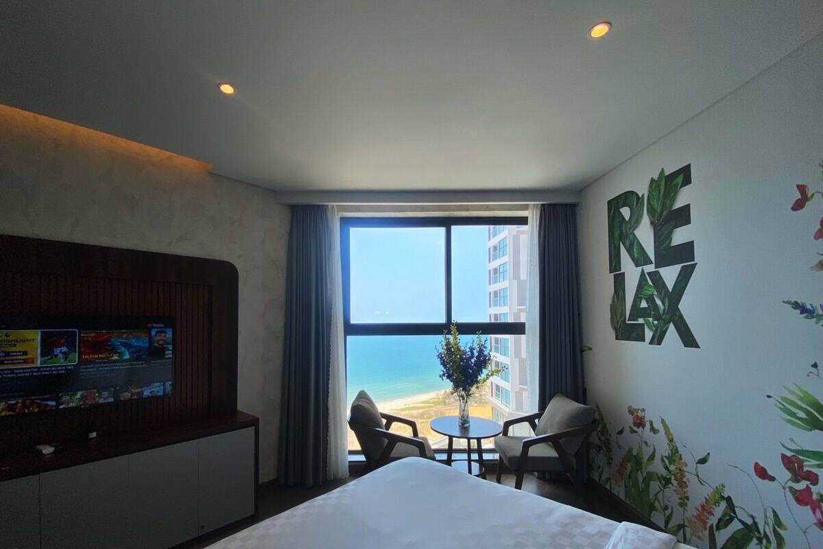 Ein Hotelzimmer mit Bett, Fernseher an der Wand, Fenster mit Blick auf den Strand und einer Wanddekoration mit dem Schriftzug 'RELAX'.