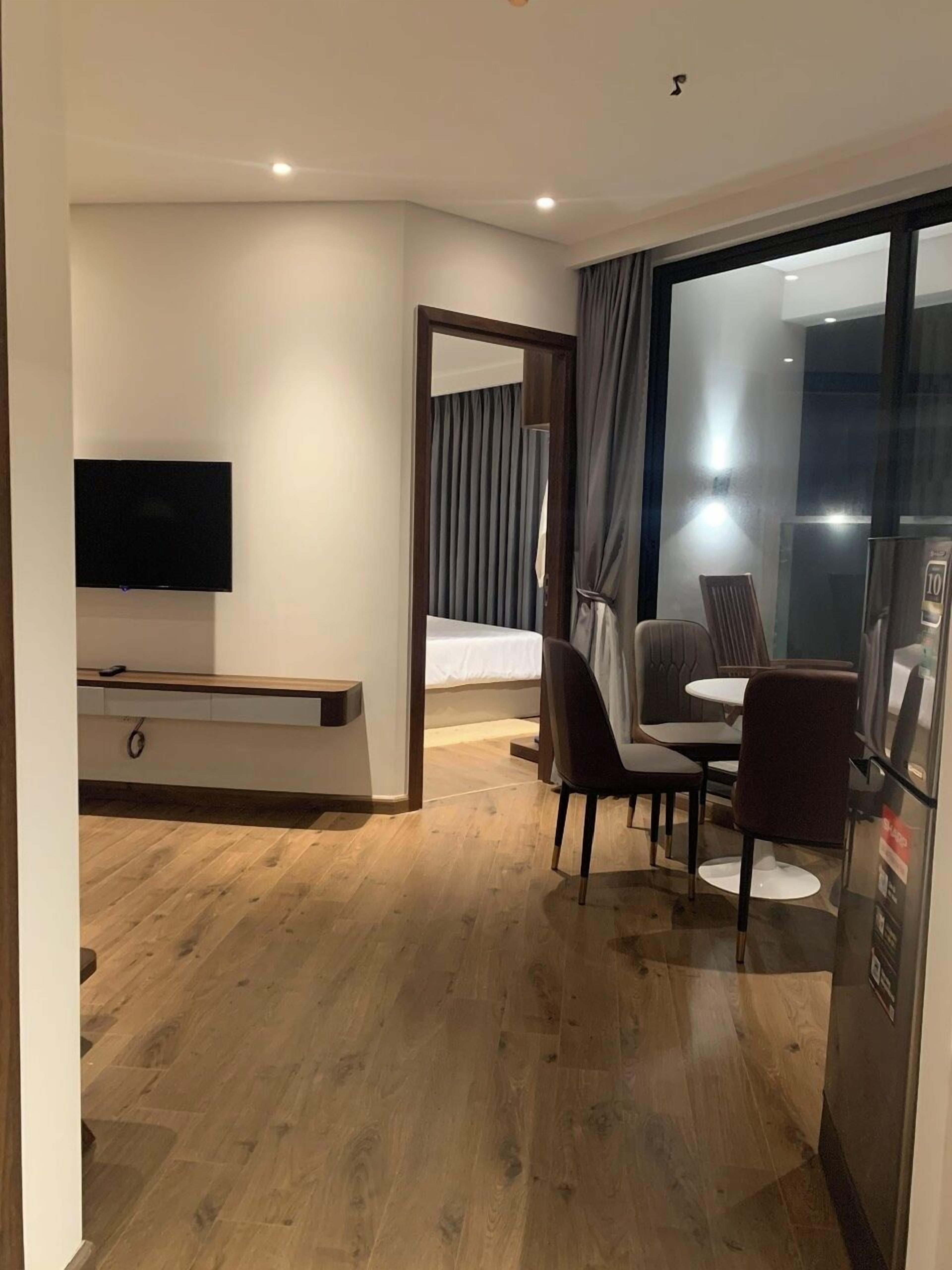 A modern hotel room with a flat-screen TV mounted on the wall, a wooden floor, a dining area with a table and chairs, and a large window with curtains.