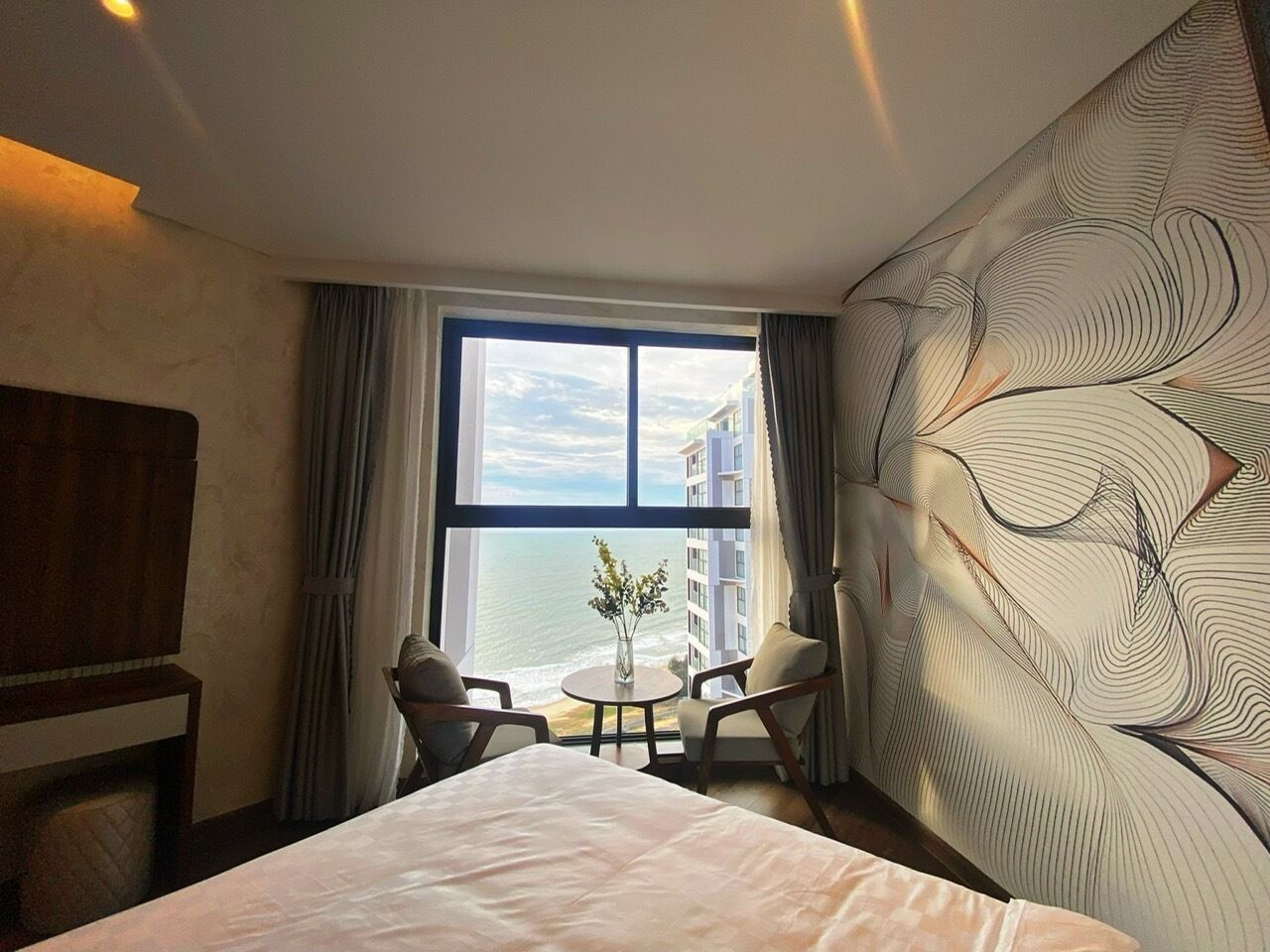 Habitación de hotel con una cama grande, dos sillones, una mesita redonda y vistas al océano a través de un ventanal.