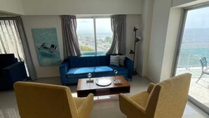 A modern living room with a blue sofa, two yellow armchairs, a coffee table, and a large window offering a view of the sea.
