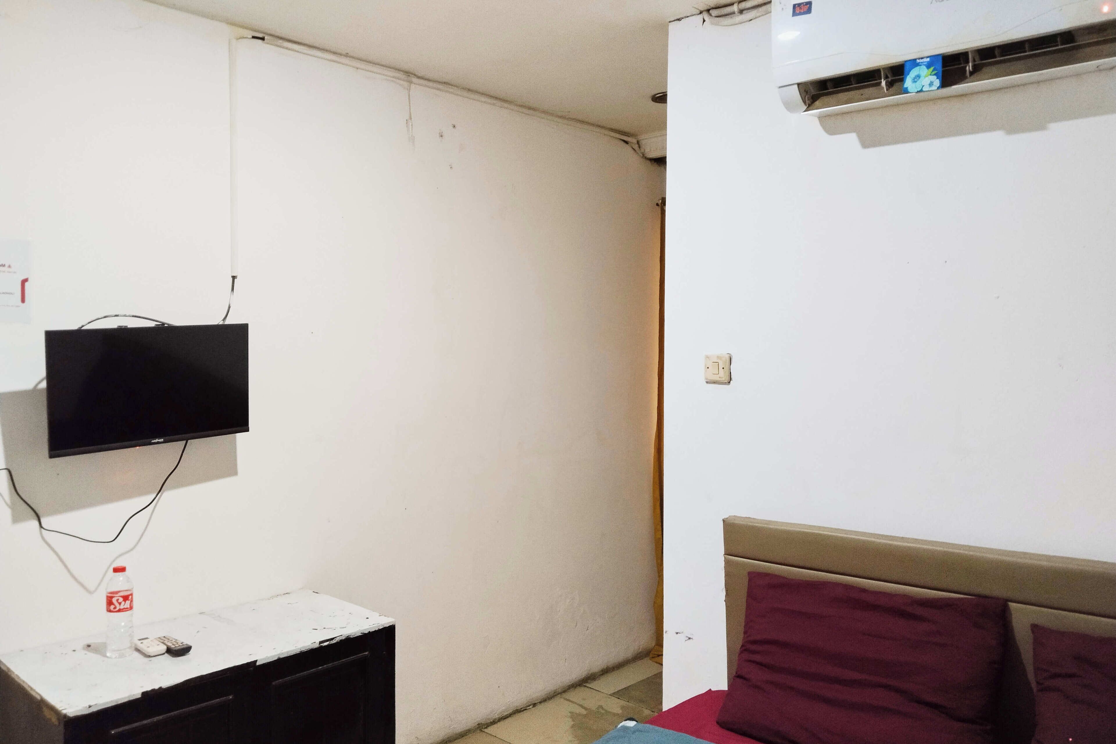 A room with a bed, a TV mounted on the wall, a small table with a bottle and remote control, and an air conditioning unit.