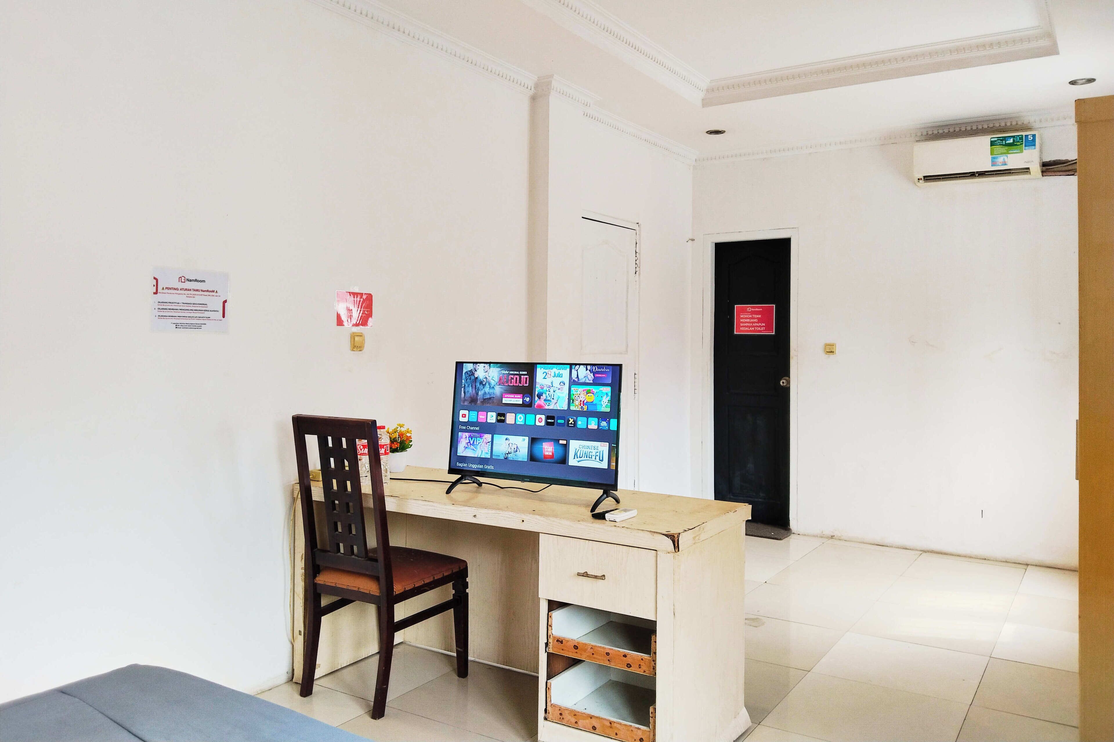 Una habitación con escritorio, silla y monitor de computadora mostrando diversas aplicaciones. Hay una puerta con un letrero, un aire acondicionado instalado en la pared y piso de baldosas.