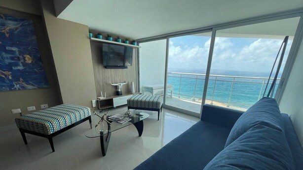 A modern living room with a sofa, a coffee table, and a TV. There is a balcony with a view of the sea.