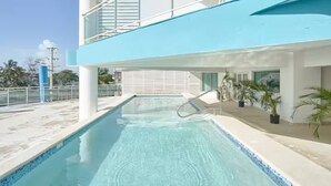 A swimming pool with a clear blue water, surrounded by a white pool deck and a blue building with a balcony.
