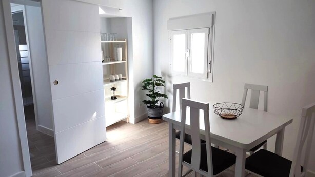 Un coin repas avec une table et des chaises blanches, une plante en pot et une étagère contenant de la verrerie et des ustensiles de cuisine.