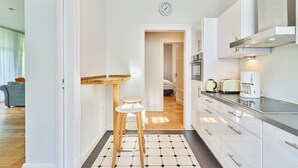 Una cocina moderna con armarios blancos, horno empotrado y cafetera. Hay un taburete de madera y una pequeña mesa con una lámpara colgante.