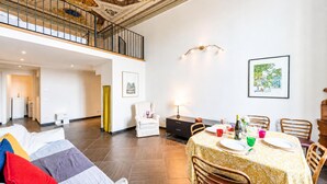 Un vaste espace de vie doté d’un haut plafond, une table à manger pour quatre personnes et un escalier menant à un niveau supérieur.
