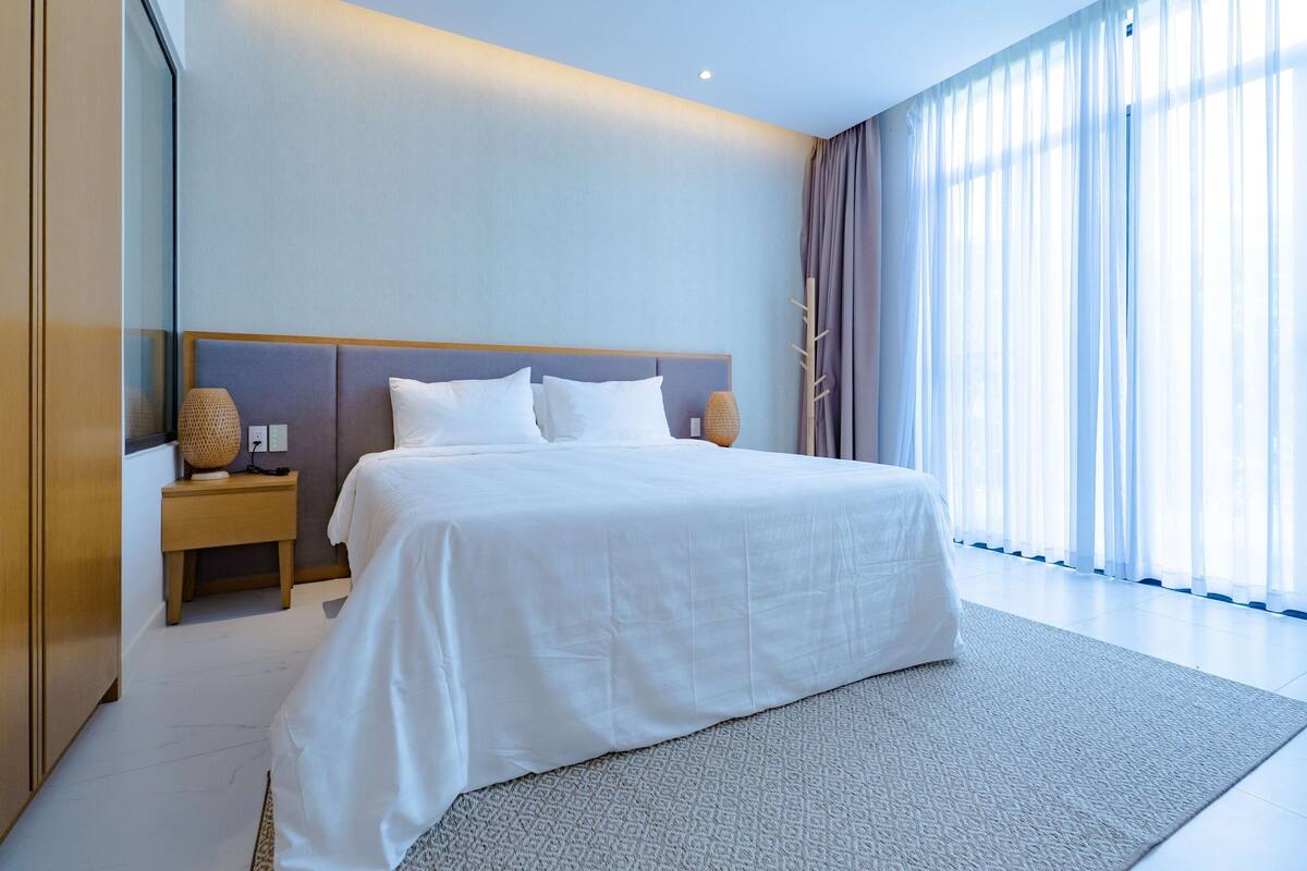 A modern hotel room with a large bed, a bedside table with a lamp, and a view of the outdoors through sheer curtains.