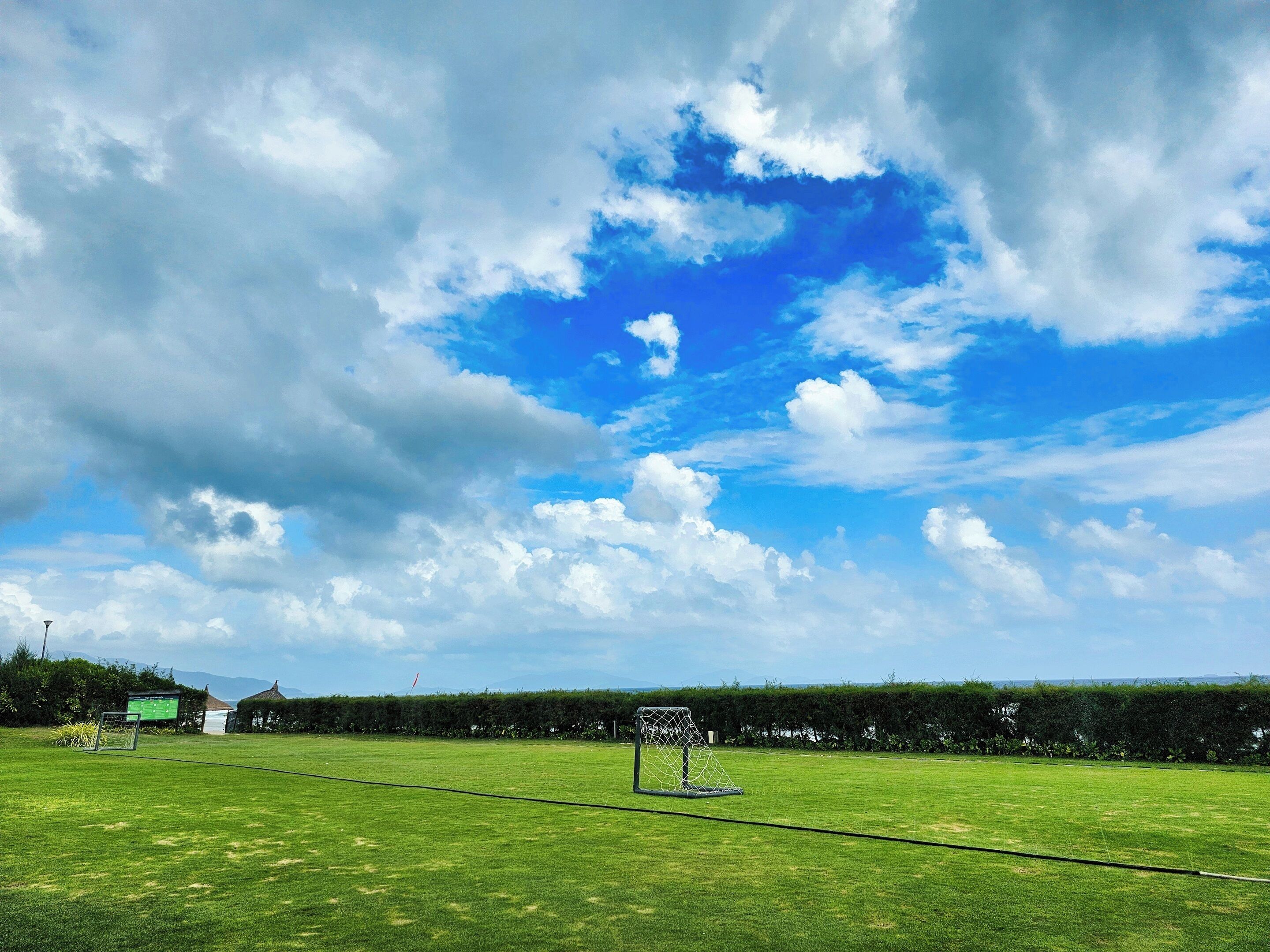 A grassy field with a soccer goal, a scoreboard, and a thatched structure in the background.