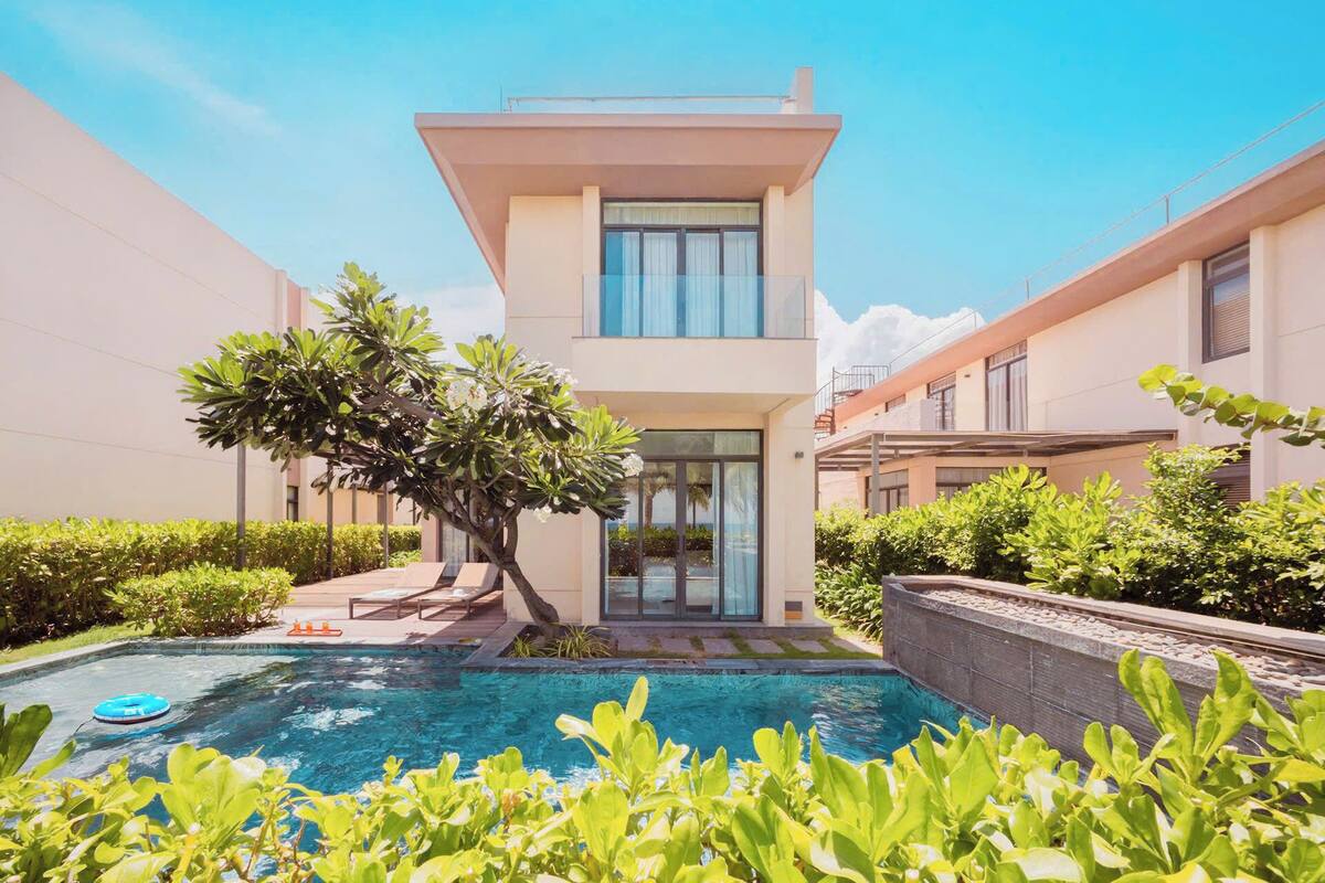 A modern two-story building with a swimming pool, surrounded by lush greenery and a clear blue sky.