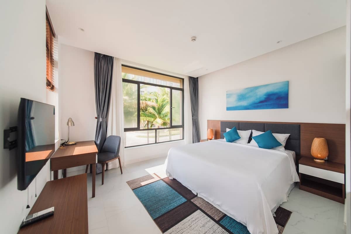 A hotel room with a bed, a desk, a chair, a TV, and a window with a view of palm trees.