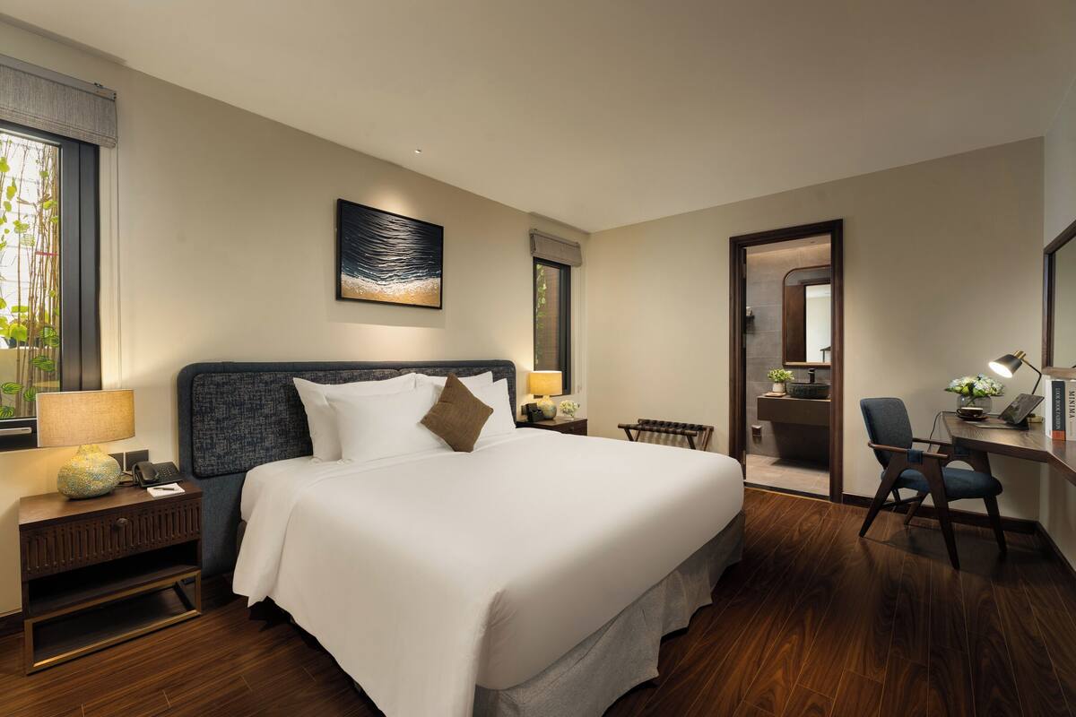 Een hotelkamer met een groot bed, een bureau met stoel, een nachtkastje en een raam met uitzicht op groen.