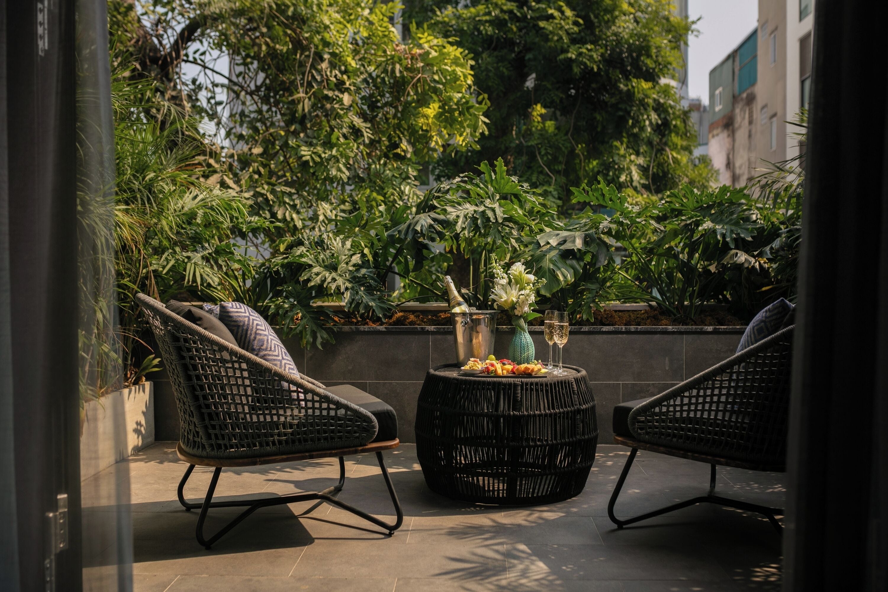 Une terrasse dotée de chaises en osier et d’une table ronde, entourée d’une végétation luxuriante et avec une vue sur la ville en arrière-plan.