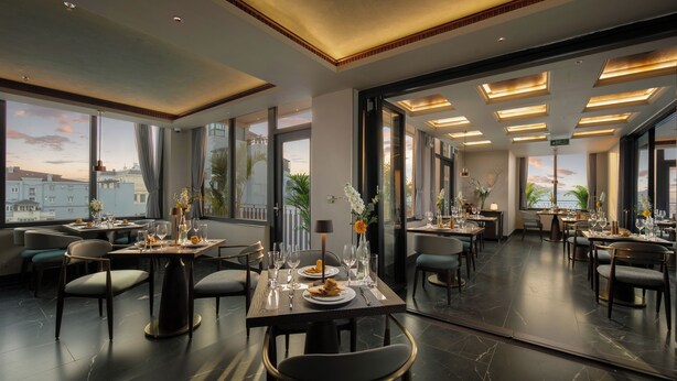 Un'elegante sala da pranzo moderna con diversi tavoli apparecchiati, ampie finestre che offrono vista sulla città e un design interno lineare e contemporaneo.