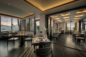 Un espace de restauration moderne, avec de nombreuses tables dressées, de grandes fenêtres offrant une vue sur le paysage urbain et un aménagement intérieur élégant et contemporain.