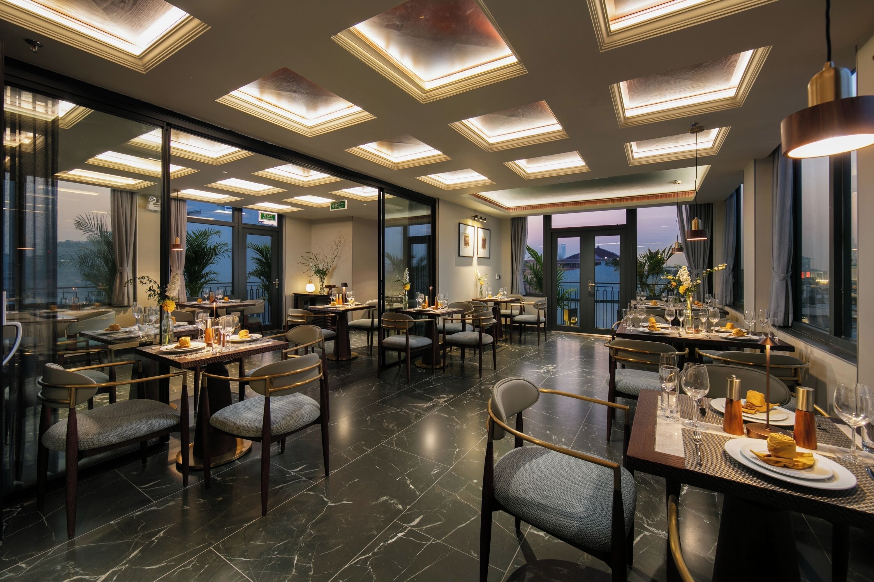A modern dining area with multiple tables and chairs, large windows, and a ceiling with recessed lighting.