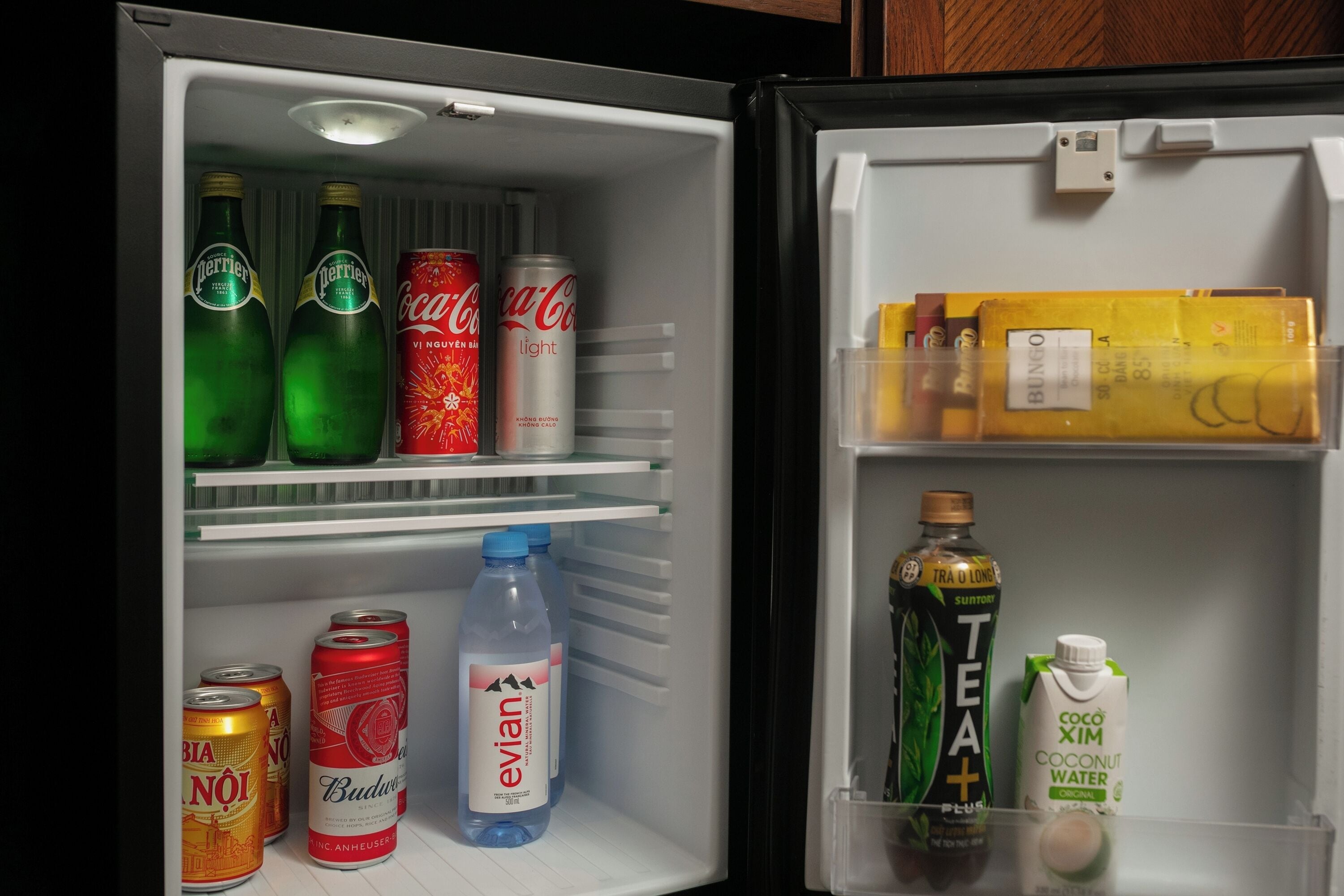 A refrigerator stocked with various beverages including Perrier, Coca-Cola, Budweiser, Evian, Bia Noi, and Teva.