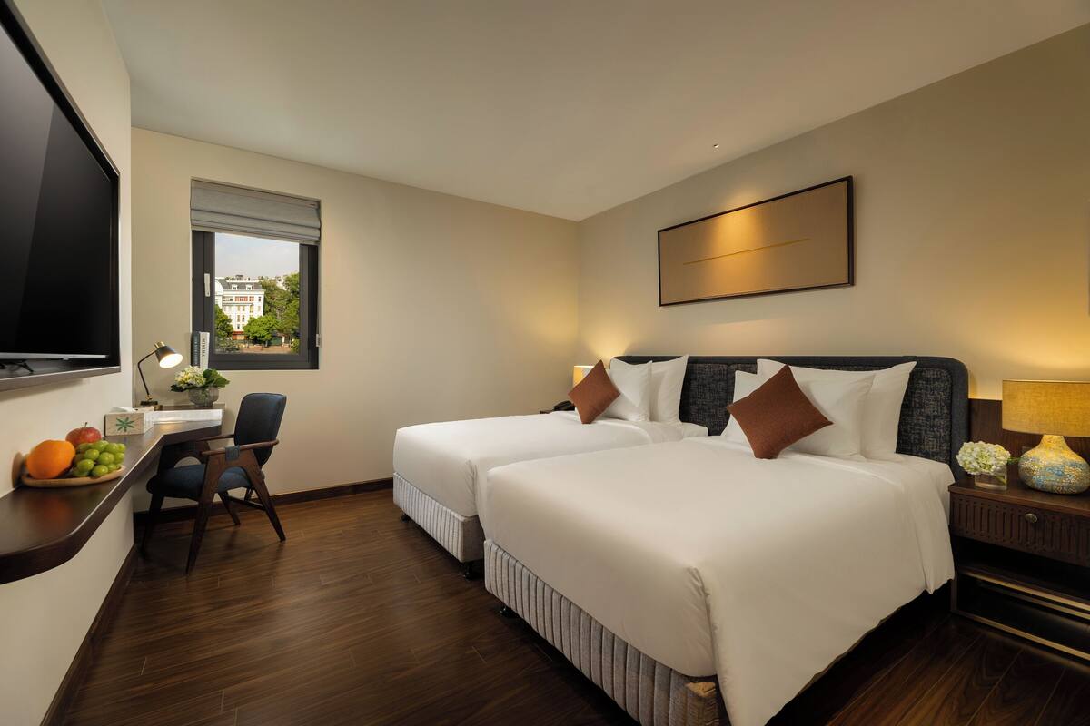 Hotelkamer met twee bedden, een bureau met stoel, een televisie en een raam met uitzicht op gebouwen.