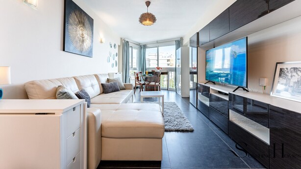 A modern living room with a sofa, a dining area, and a large TV mounted on the wall. - Stylish Retreat: 1-Bed, Pool, Sleeps 3 REF001 (Benalmádena)