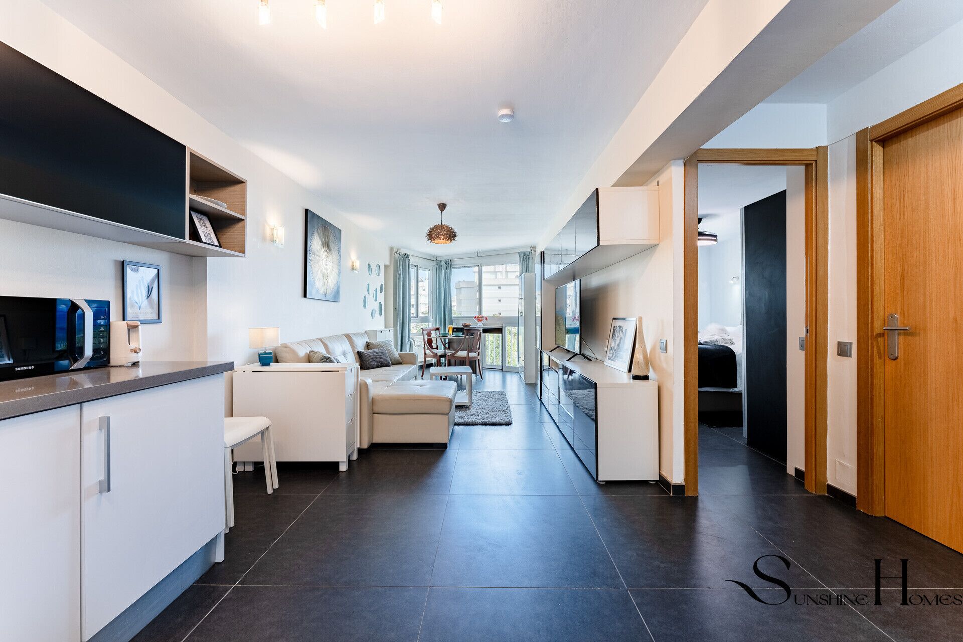Una cocina moderna con microondas, tostadora y armarios. Hay un comedor con mesa y sillas, y una sala de estar con sofá y televisor. La habitación tiene un ventanal grande y una puerta que conduce a otra estancia.