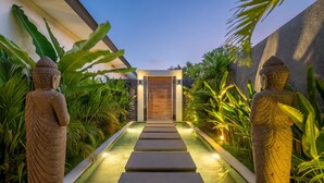 A pathway leading to a modern building entrance, flanked by large stone statues and lush tropical plants. - Private Pool • Outdoor Showers • Near Beaches (Uluwatu)