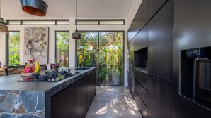A modern kitchen with dark cabinetry, a marble countertop, and a large window overlooking a tropical garden. - Private Pool • Outdoor Showers • Near Beaches (Uluwatu)
