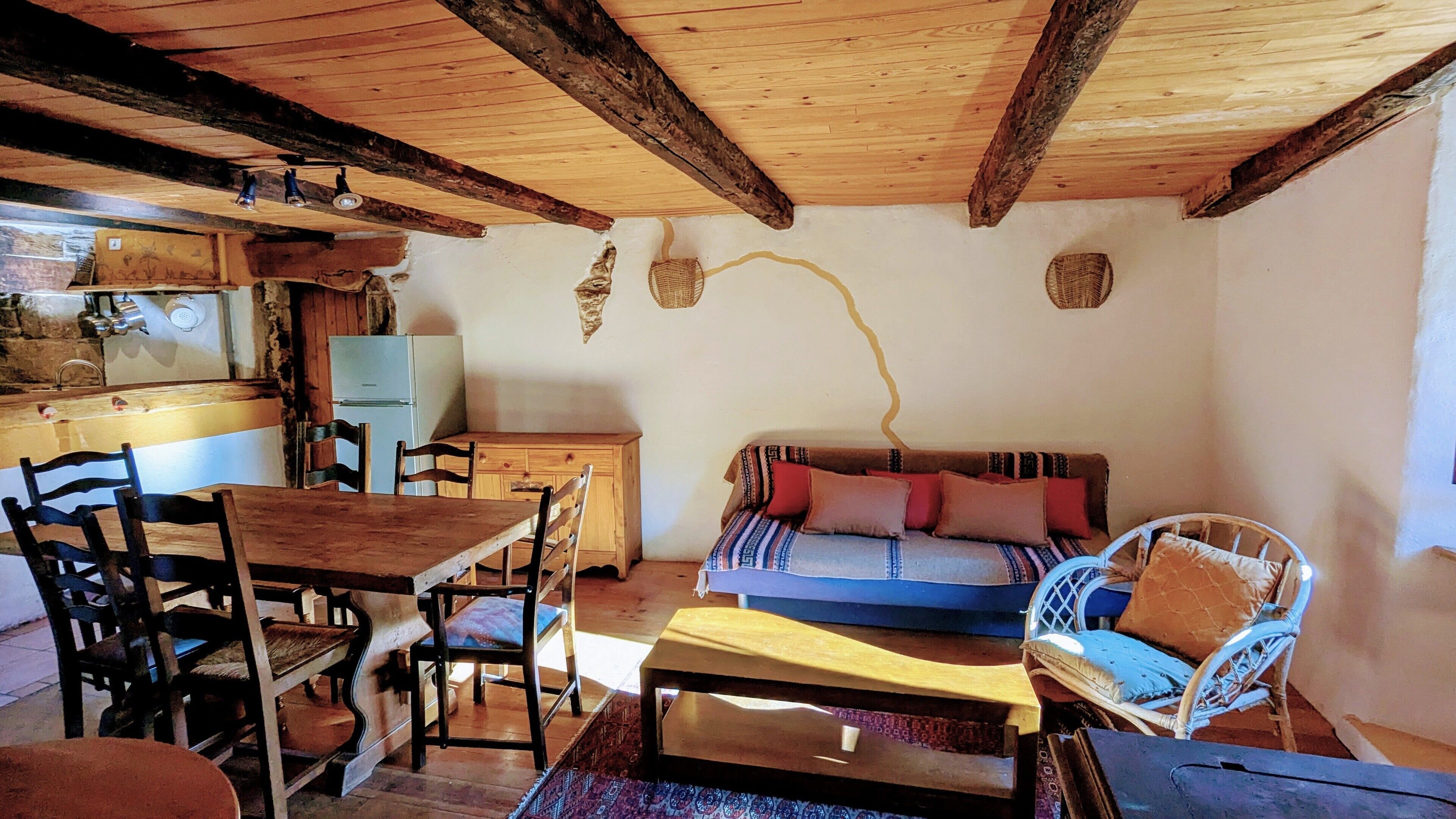 A rustic interior with exposed wooden beams, a dining area with wooden chairs, a sofa with patterned cushions, and a wicker chair.