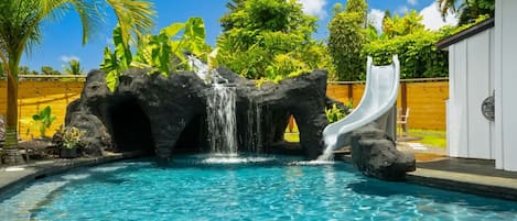 Ein Pool mit Wasserfall und Rutsche, umgeben von tropischer Vegetation und einem Holzzaun.