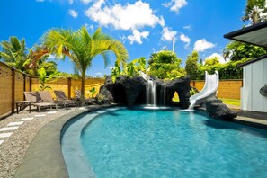 A swimming pool with a waterfall and slide, surrounded by lounge chairs and palm trees.