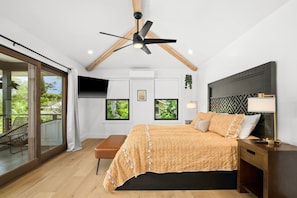 A bedroom with a bed, a ceiling fan, a TV, and a balcony with a chair.