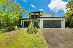 A modern two-story house with a large garage, surrounded by lush greenery and palm trees.