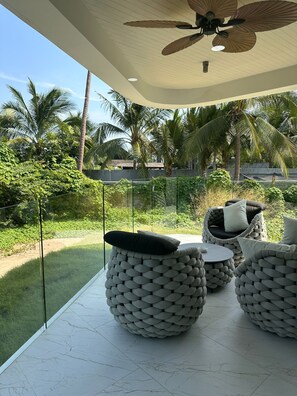 A modern outdoor seating area with round, woven furniture and a glass railing overlooking a lush garden with palm trees. - Beachside Residence (Koh Samui)