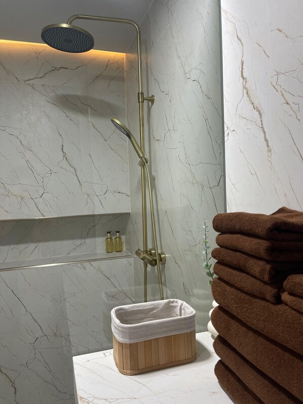 A bathroom with a marble wall, a shower with a golden fixture, a wooden bath mat, and a stack of towels. - Beachside Residence (Koh Samui)