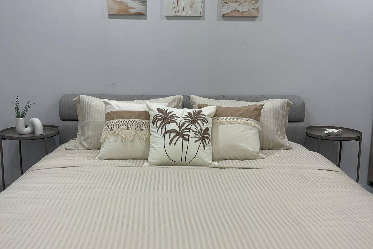 Una cama bien hecha con una colcha a rayas y almohadones decorativos, incluyendo uno con diseños de palmeras. Encima de la cama, hay tres obras de arte enmarcadas en la pared.