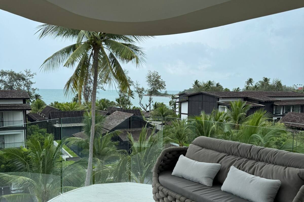 Un espacio moderno al aire libre con una mesa redonda y un sofá, con vistas a un paisaje costero con palmeras y casas.