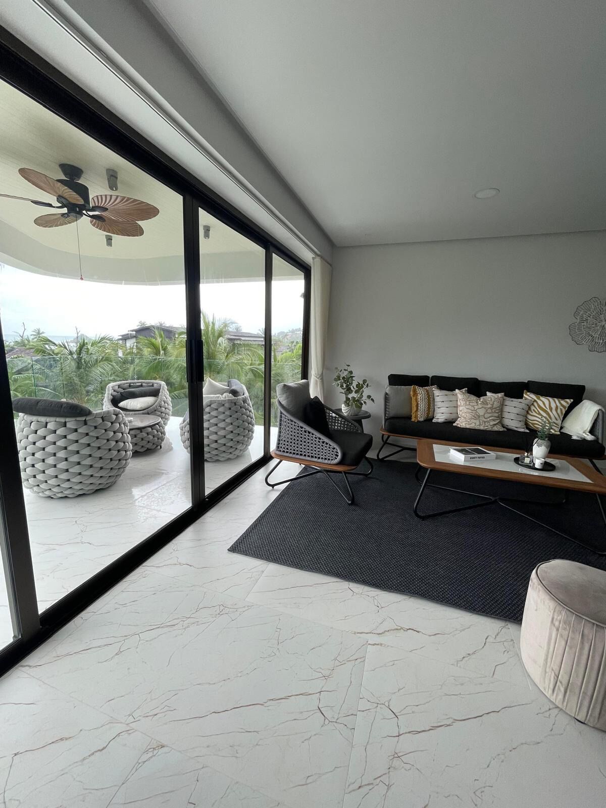 A modern living room with a large sliding glass door leading to a balcony, a black sofa with patterned cushions, and a coffee table with a book and a small plant.