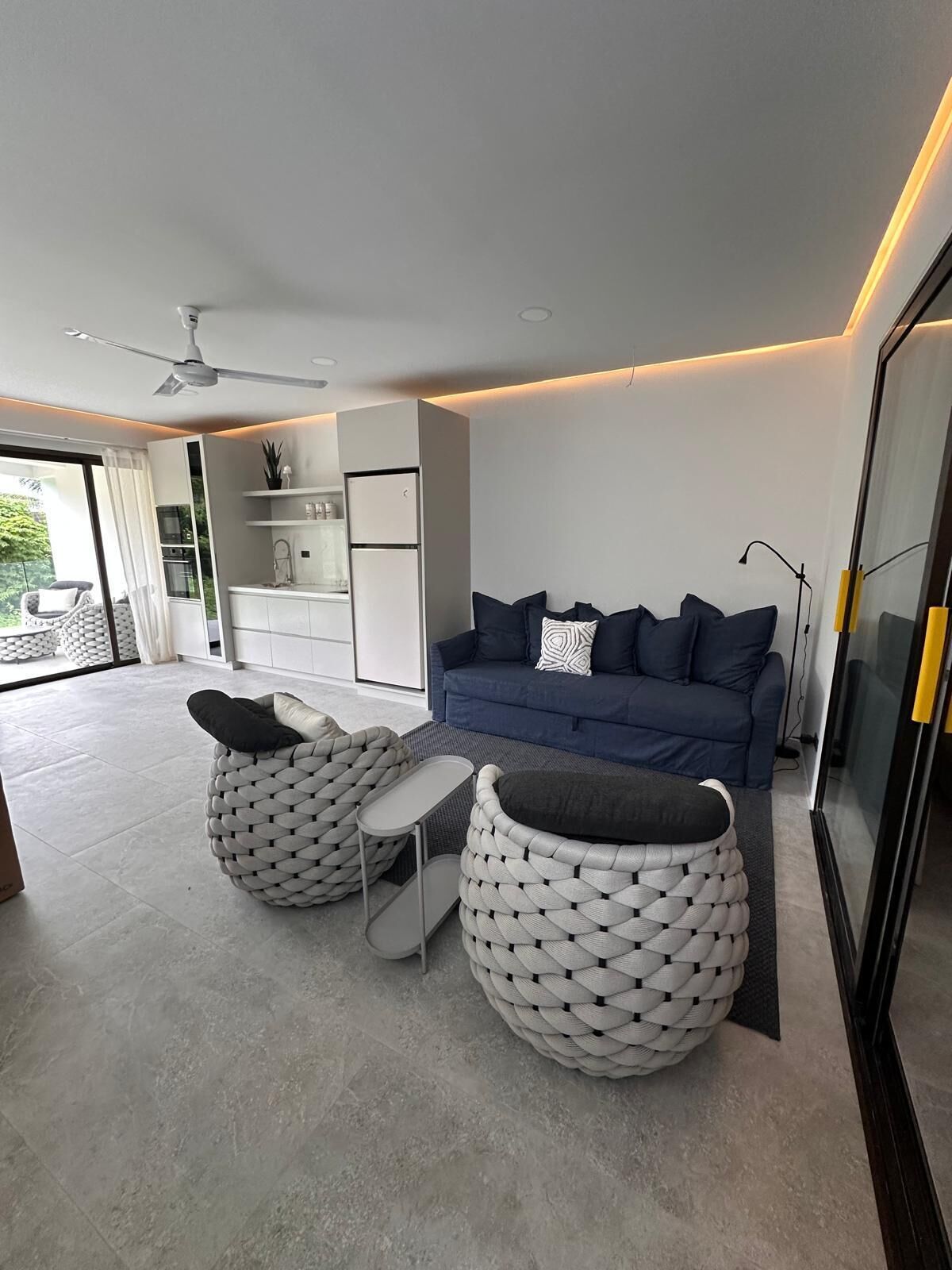 A modern living room with a blue sofa, two white pouf chairs, a small table, and a ceiling fan.