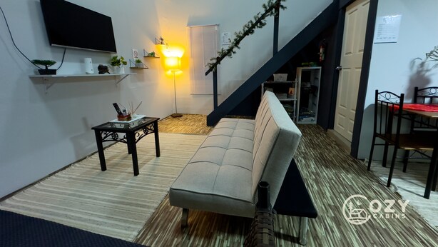 A modern living room with a sofa, a small table, a mounted TV, and a staircase leading to another room. - Cozy Cabin • 5 mins from Cave Tubing & Zipline (Frank's Eddy)