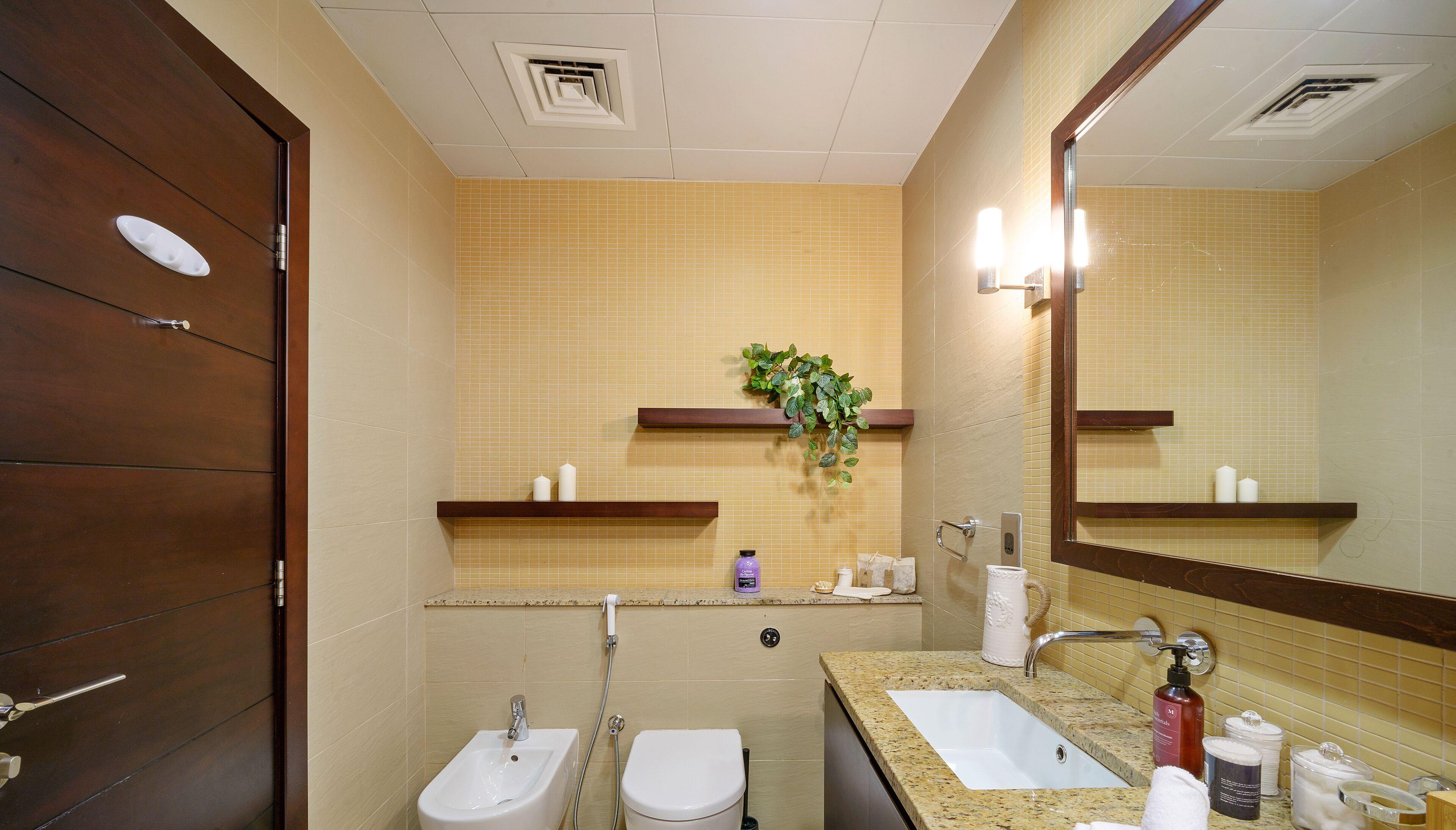 A bathroom with a wooden door, a sink, a toilet, a mirror, and a shelf with toiletries.