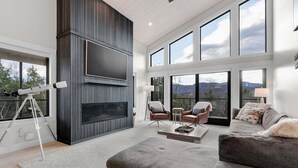 Ein modernes Wohnzimmer mit großem Fernseher, bequemen Sitzmöglichkeiten und großen Fenstern mit Blick auf die Berge.